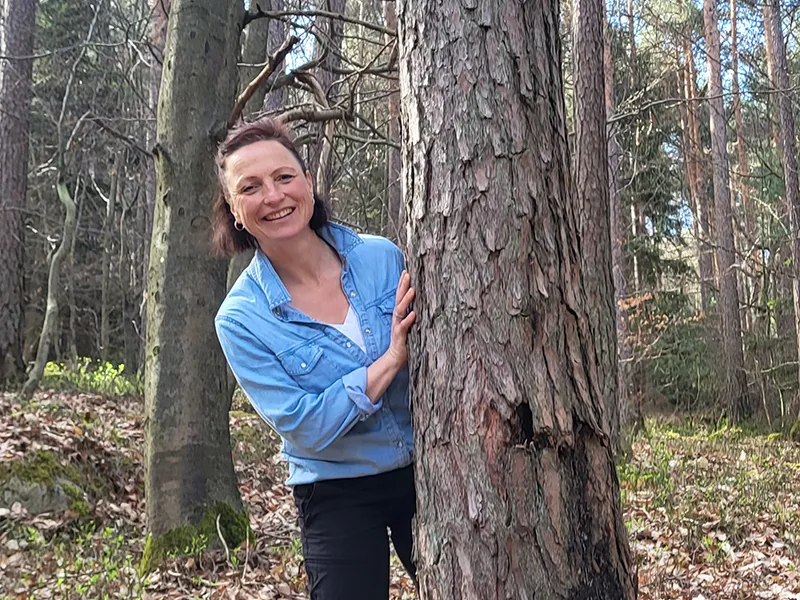 Waldpädagogin Gabi steht im Wald hinter einem Baum und kuckt hervor