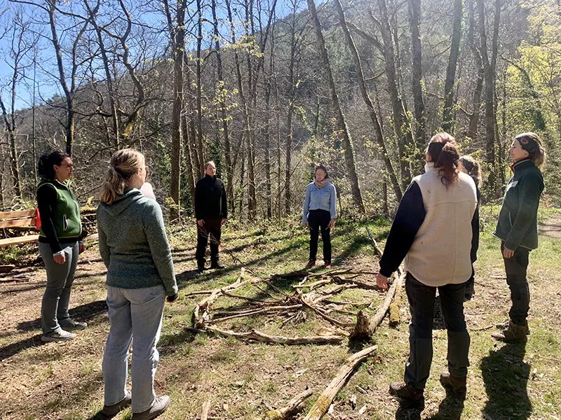 Erwachsene Frauen stehen im Wald um einen aus Holz gelegten Kreis, sie haben die Augen geschlossen.